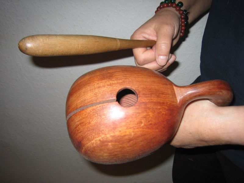 A hand holds a rounded wooden moktak with a narrow slit and a handle; a separate wooden beater is held above it